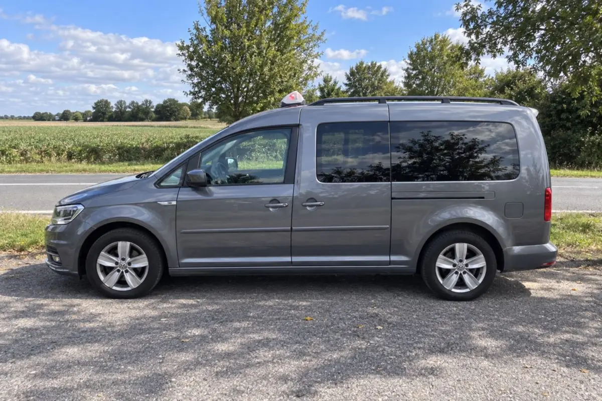 Le taxi JB TRANSPORT vu de profil : un Volkswagen Caddy Maxi gris aux vitres arrière teintées, au bord d’une route de campagne